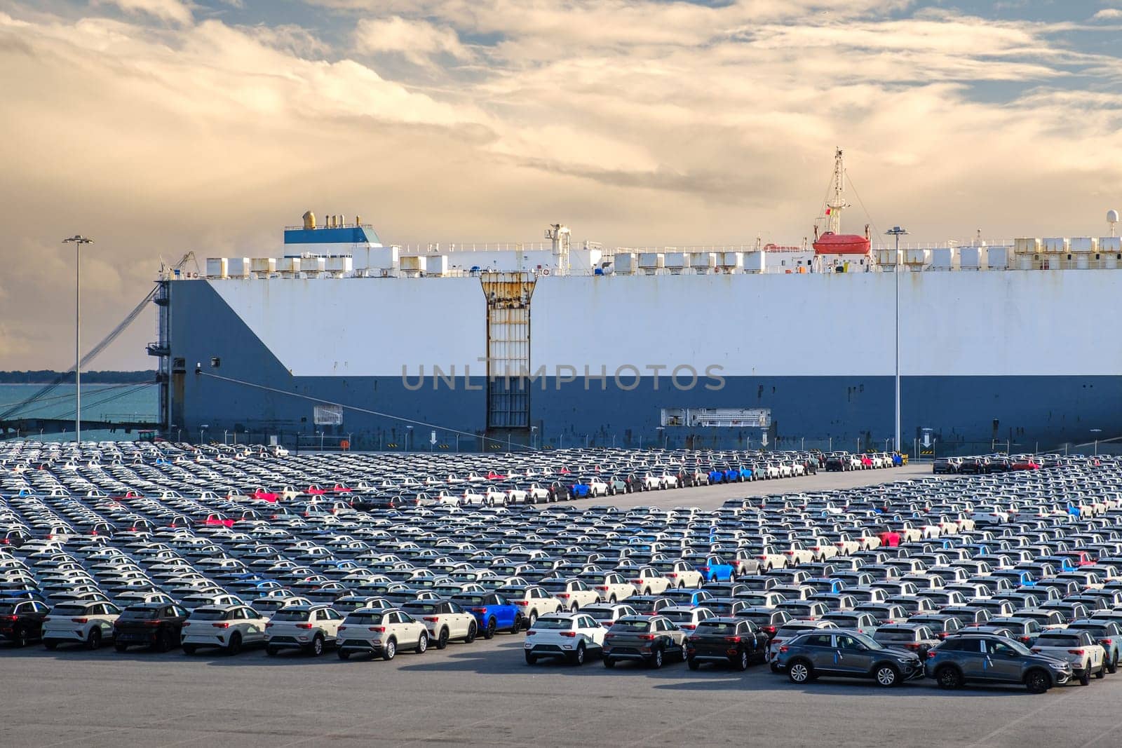 Large vehicle carrier ship known as RO-RO at port on sunny day, large parking area filled with cars waiting loading and transportation to buyers and car dealers. Blue sky and colorful clouds, vehicle transportation.
