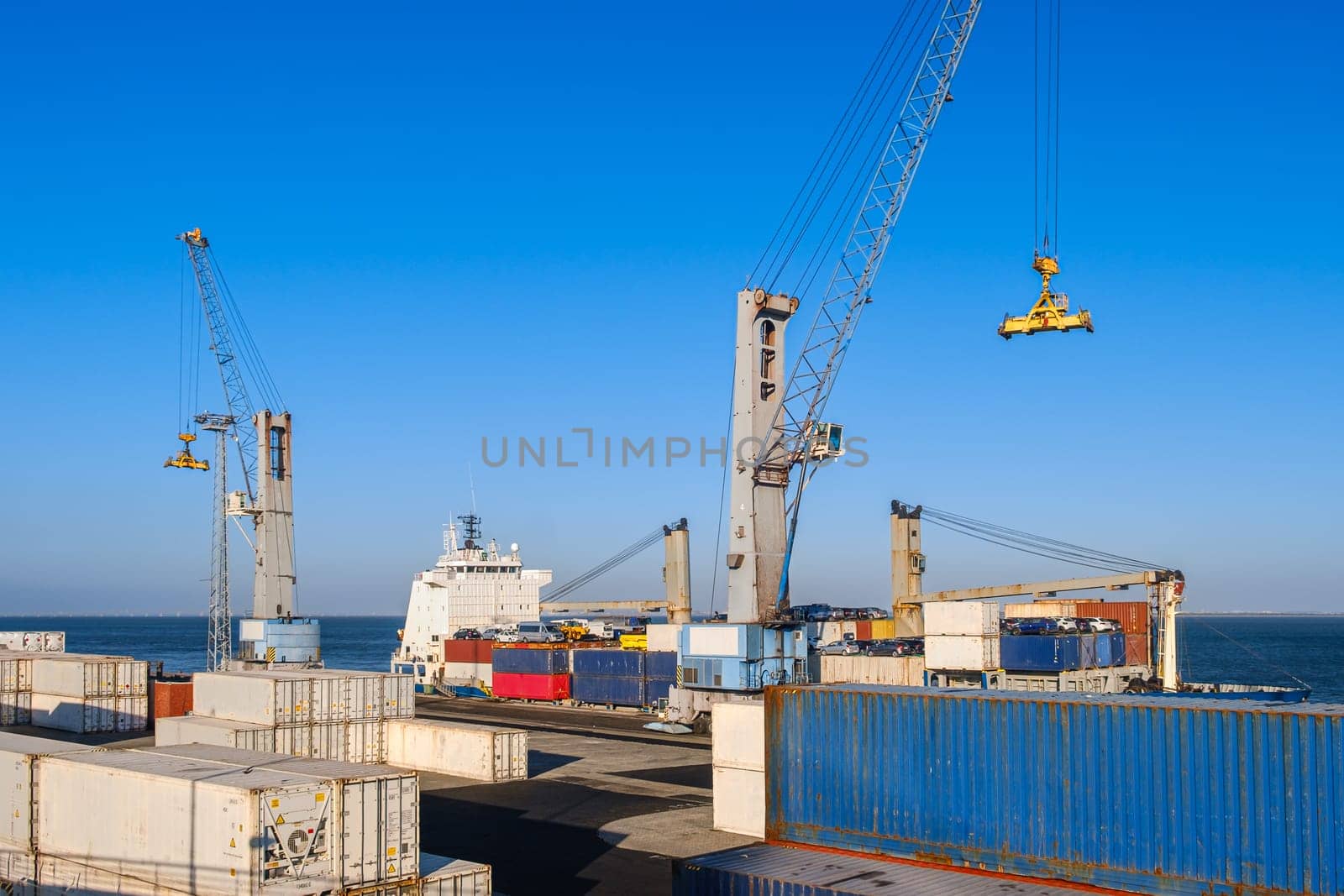 Maritime logistics and transportation scene at commercial seaport showing stacked shipping containers in various colors, cargo handling equipment, and vessel cranes against clear blue sky background. Cars visible on dock.