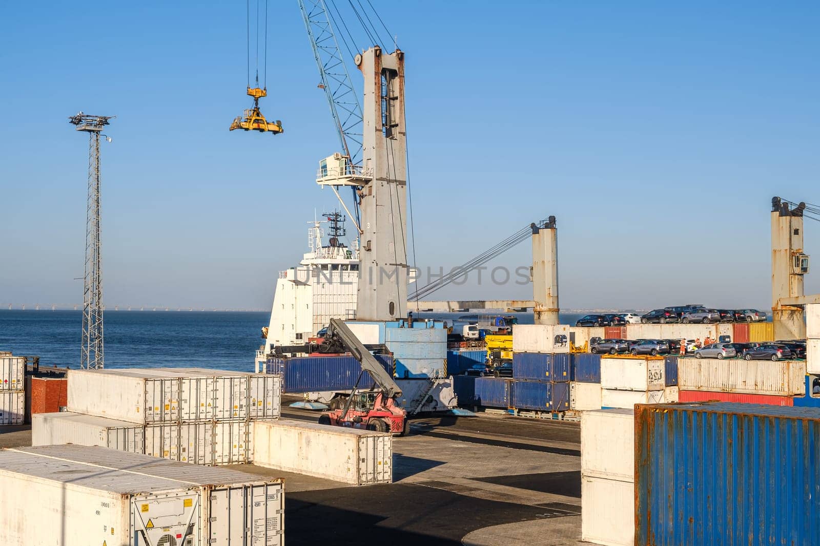 Maritime logistics and transportation scene at commercial seaport showing stacked shipping containers in various colors, cargo handling equipment, and vessel cranes against clear blue sky background. Cars visible on dock.