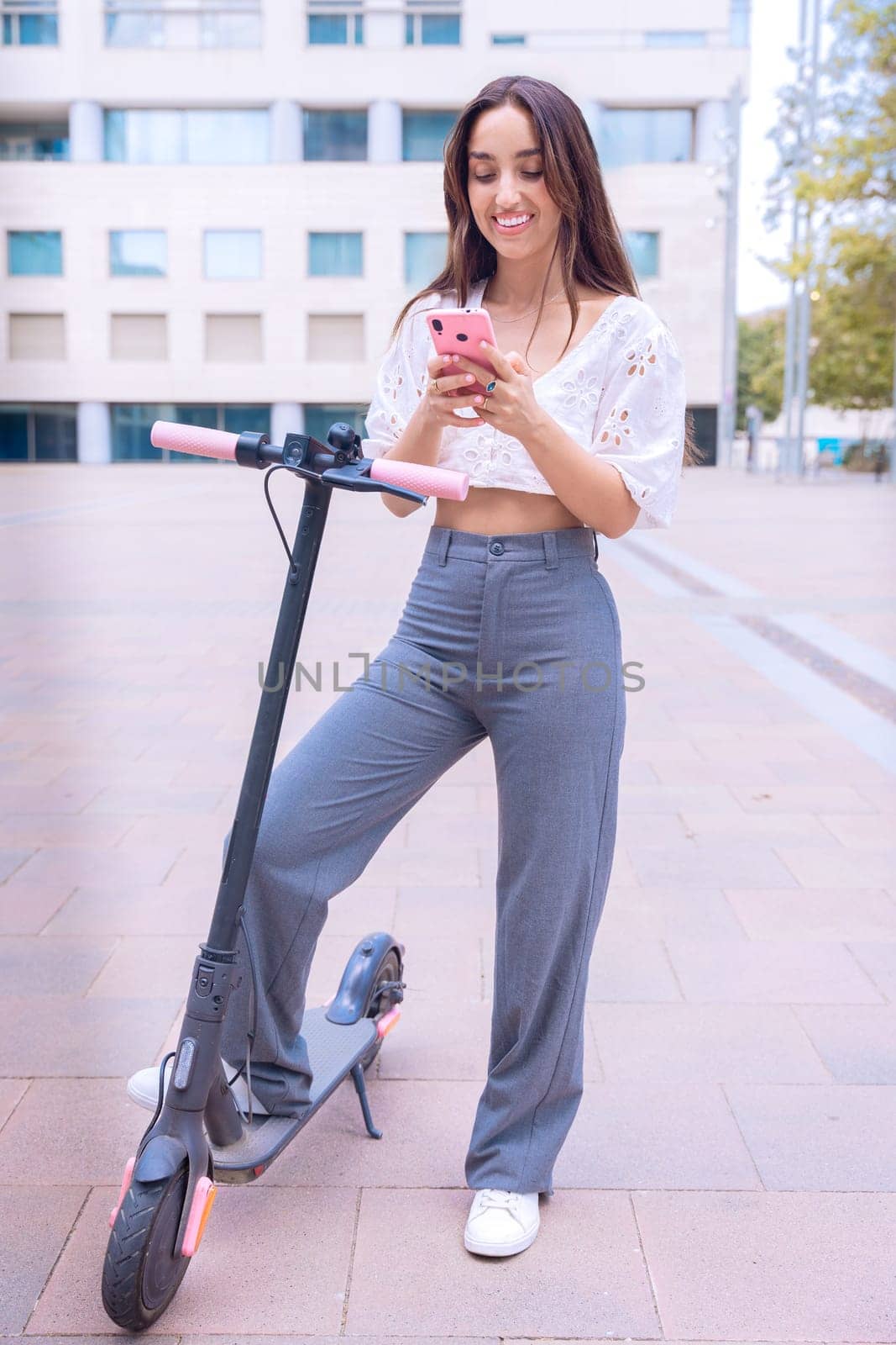 smiling young woman in a stylish white top uses her smartphone while standing next to an electric scooter in an urban setting. She appears excited and engaged, enjoying modern technology and mobility.