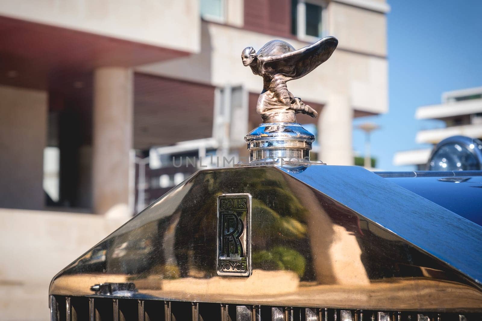 Lisbon, Portugal - Sep 29, 2024: Close-up and selective focus on the Rolls-Royce mascot hood ornament or bonnet badge called Spirit of Ecstasy, female in billowing cloth like wings behind, leaning forward, and emblem on grille. Rare kneeling version