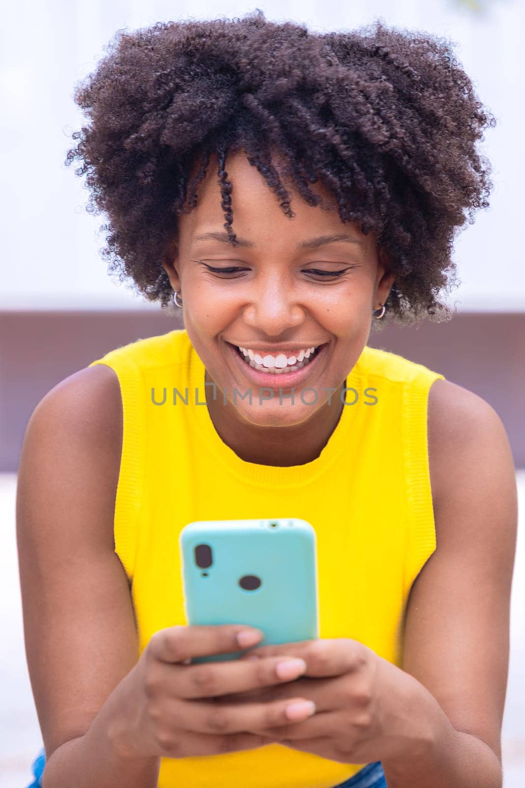 A young woman with natural curly hair wearing a yellow top rests her face on her hand while holding a smartphone. Calm and happy moment captured in a bright outdoor setting.