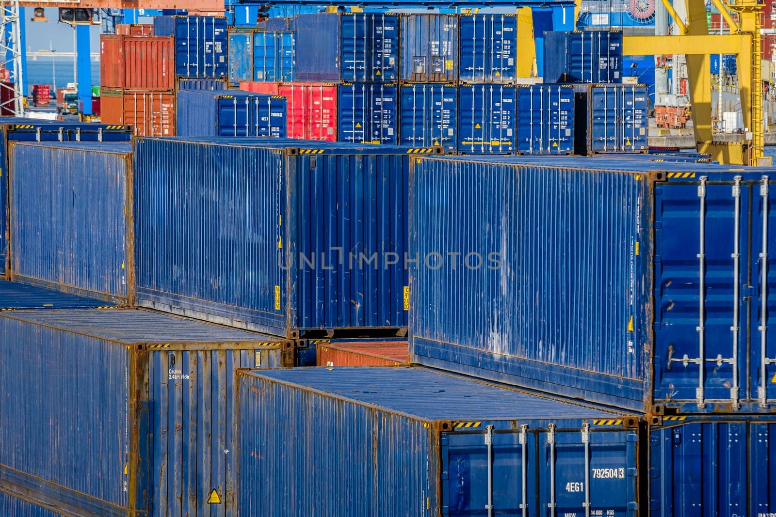 Close view of rows of blue cargo containers stacked at docking station in sunny day. Cranes in the background ready to load and unload cargo, maritime trade, shipping yard, storage, global logistics, freight.