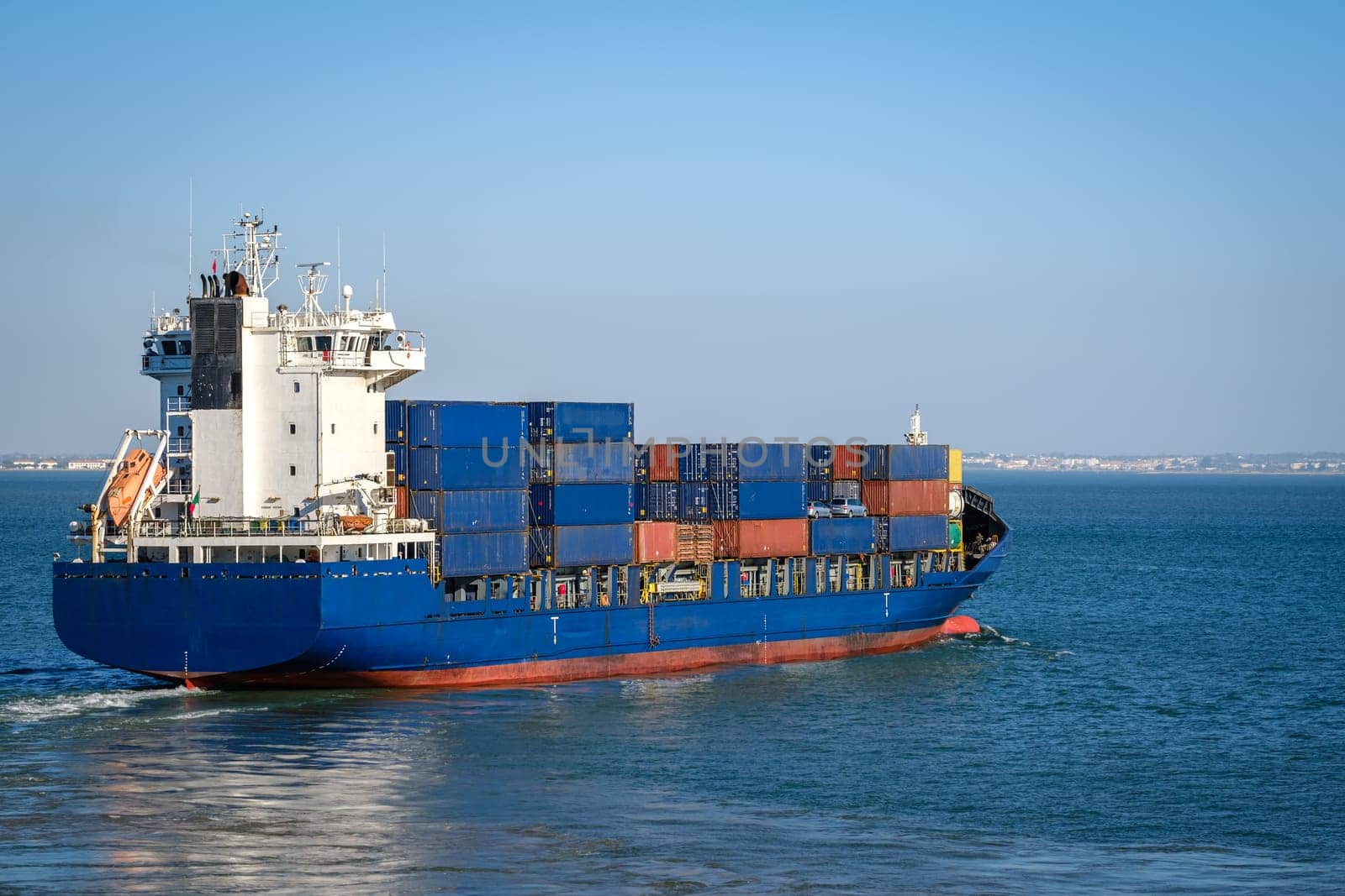 Modern blue bulk carrier loaded with stacked shipping containers navigating harbor near sea port. Global trade, maritime logistics, cargo, international freight.
