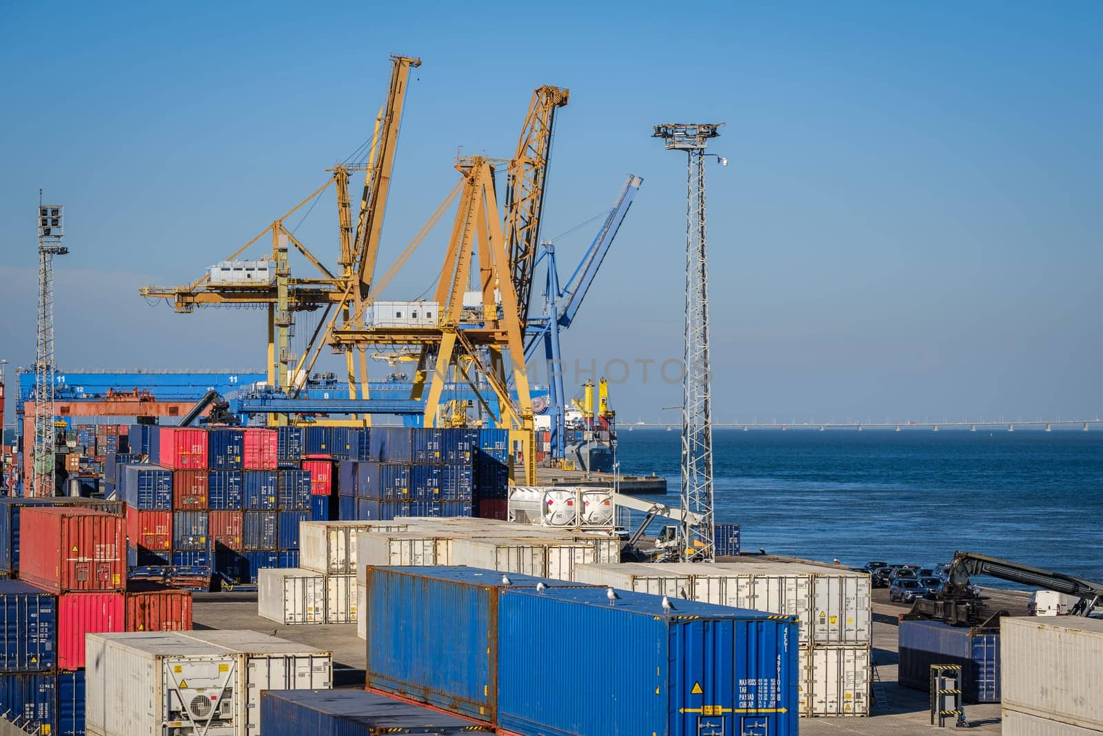 Maritime logistics and transportation scene at commercial seaport with stacked shipping containers, gantry and STS cranes, reach stackers in cargo terminal. International shipping and freight infrastructure, clear blue sky and sea waters.