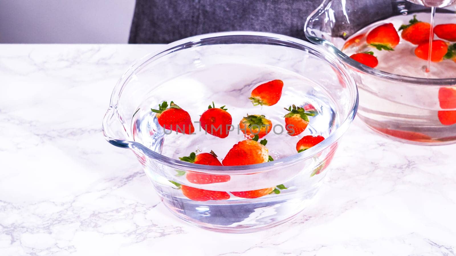 Ripe strawberries are submerged in water within a large glass mixing bowl, a step in washing the fruit to ensure cleanliness and longevity before storage or consumption.