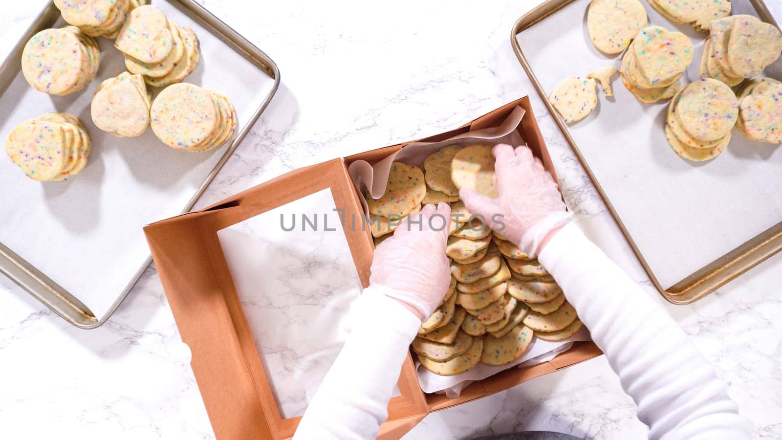 Flat lay. With precision, the woman is carefully arranging the sugar cookies, filled with dough-mixed sprinkles, into a rustic brown paper box.