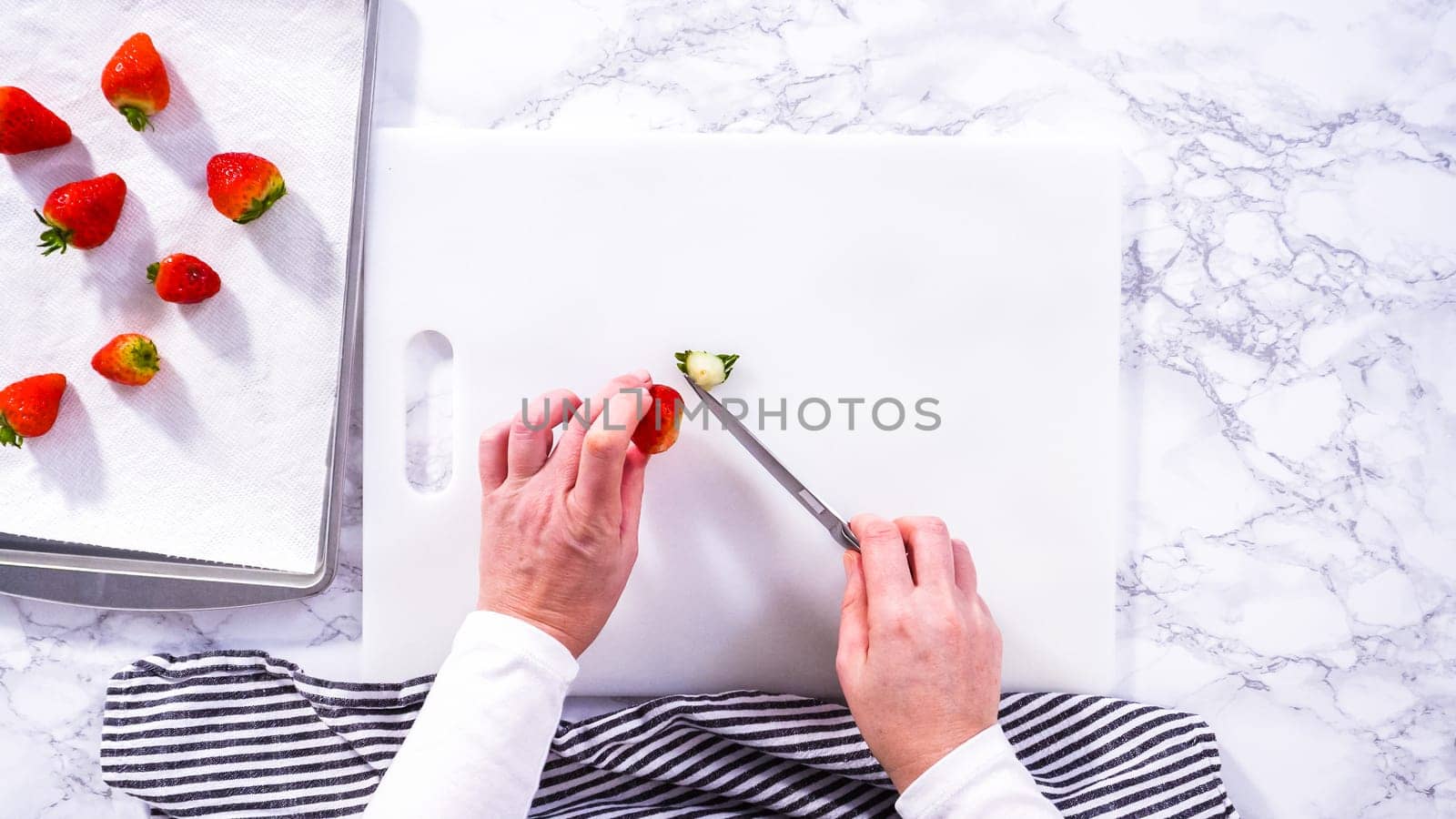 Flat lay. Succulent red strawberries, some showing early spoilage, are arrayed on a white cutting board, held securely in place, with a paper towel lined glass bowl nearby suggesting an attempt at preservation.