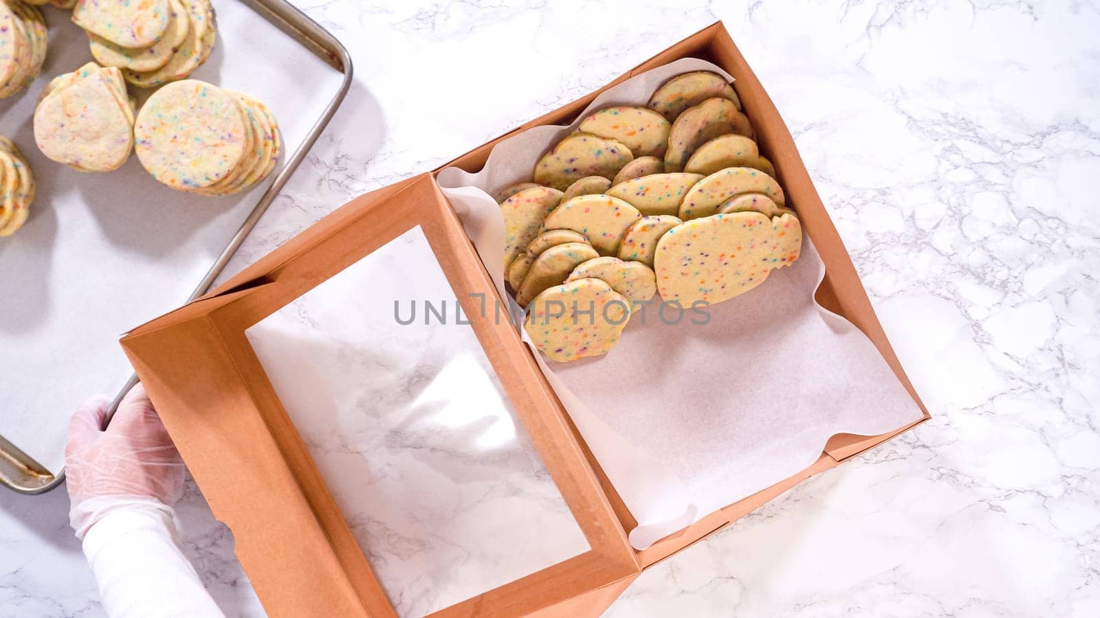 Flat lay. With precision, the woman is carefully arranging the sugar cookies, filled with dough-mixed sprinkles, into a rustic brown paper box.