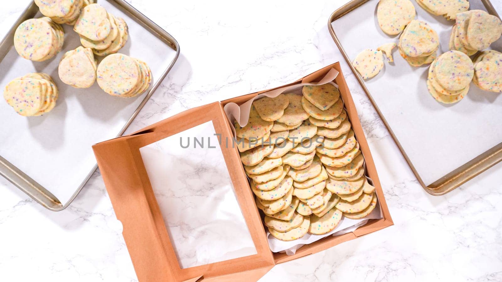 Flat lay. With precision, the woman is carefully arranging the sugar cookies, filled with dough-mixed sprinkles, into a rustic brown paper box.