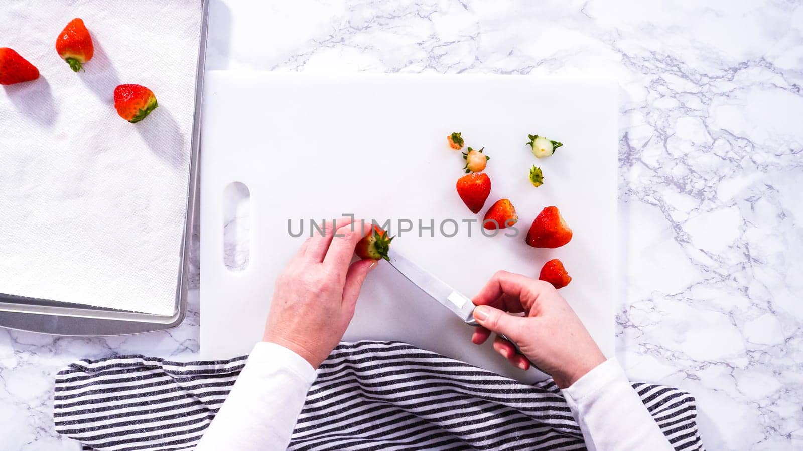 Flat lay. Succulent red strawberries, some showing early spoilage, are arrayed on a white cutting board, held securely in place, with a paper towel lined glass bowl nearby suggesting an attempt at preservation.