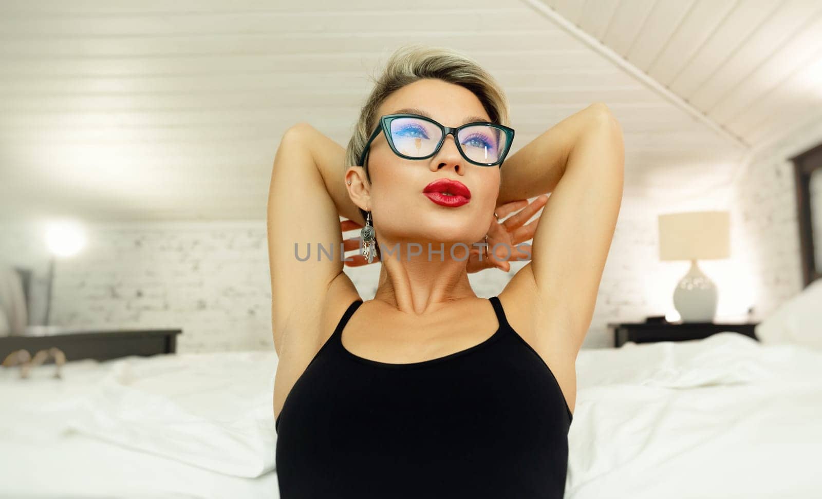 Portrait of a sexy girl with short hair and glasses, in a sexy black dress, emotionally echoing, demonstrating confidence on the bed in the bedroom