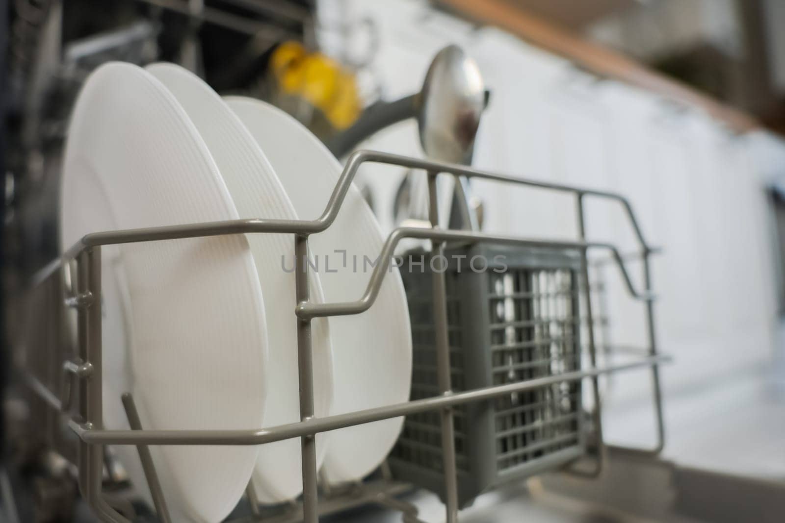 Close-up of open dishwasher department with clean plates and cutlery against kitchen background: white cabinets and wooden countertop out of focus. The concept of washing dishes, cleanliness, alleviating household chores. High quality photo