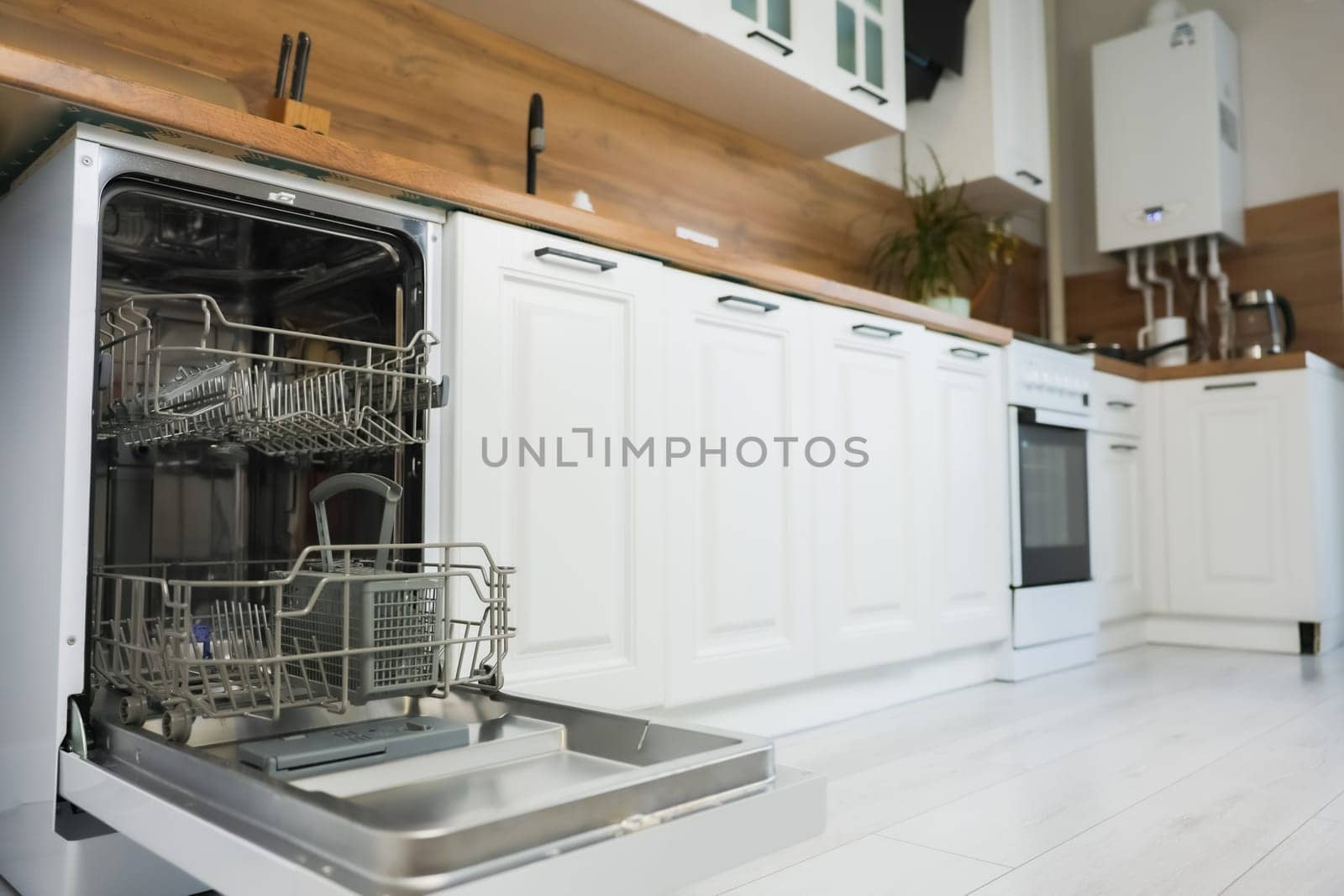 Empty dishwasher with two compartments in gray tones stands with the door open, ready to load, against the background of a white and wooden kitchen in out-of-focus. High quality photo