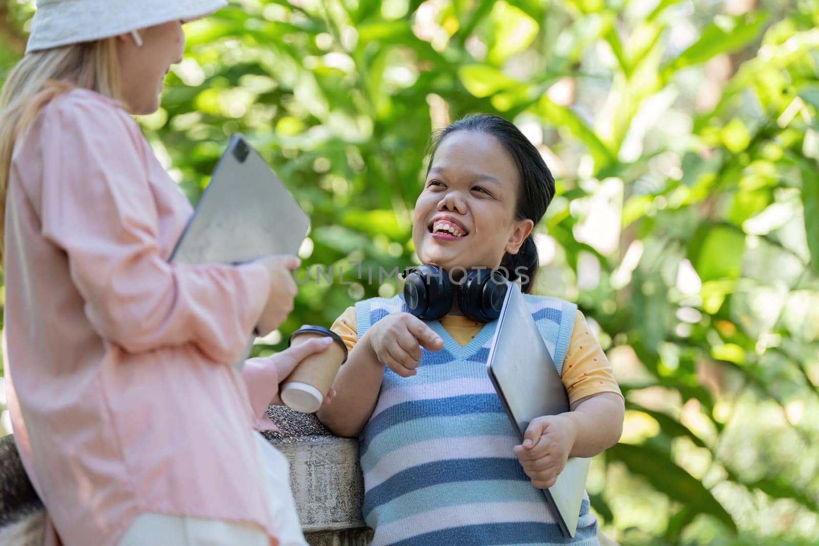 Two friends, one with achondroplasia, engaged in a lively discussion outdoors, sharing ideas and technology in a lush green park.
