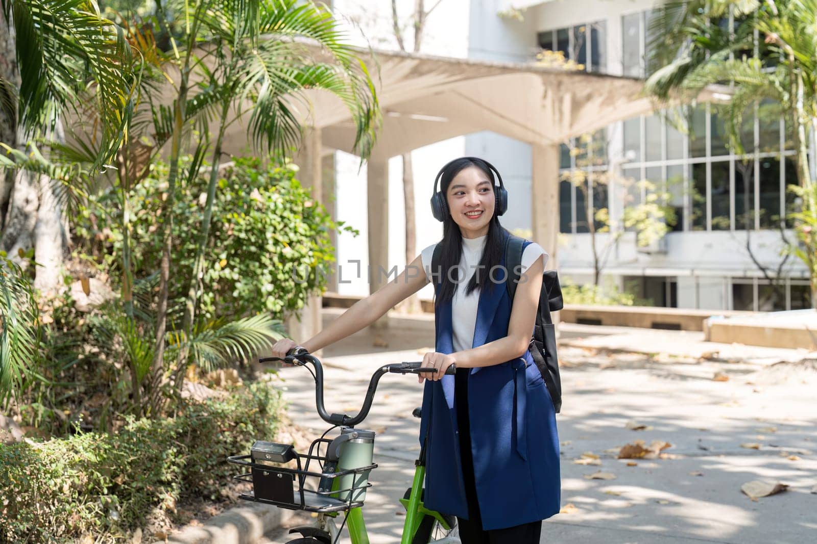 A friendly businesswoman with headphones smiles while riding her sustainable bicycle through a lush urban pathway, promoting energy saving transportation and a healthy lifestyle.