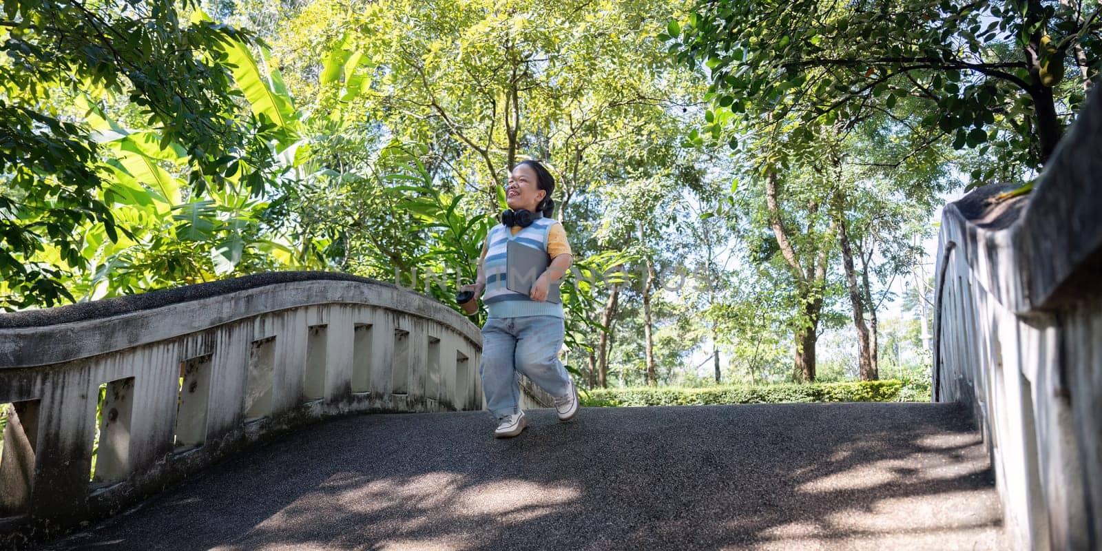A happy child with achondroplasia wearing headphones, running energetically across a bridge surrounded by vibrant greenery in a park.