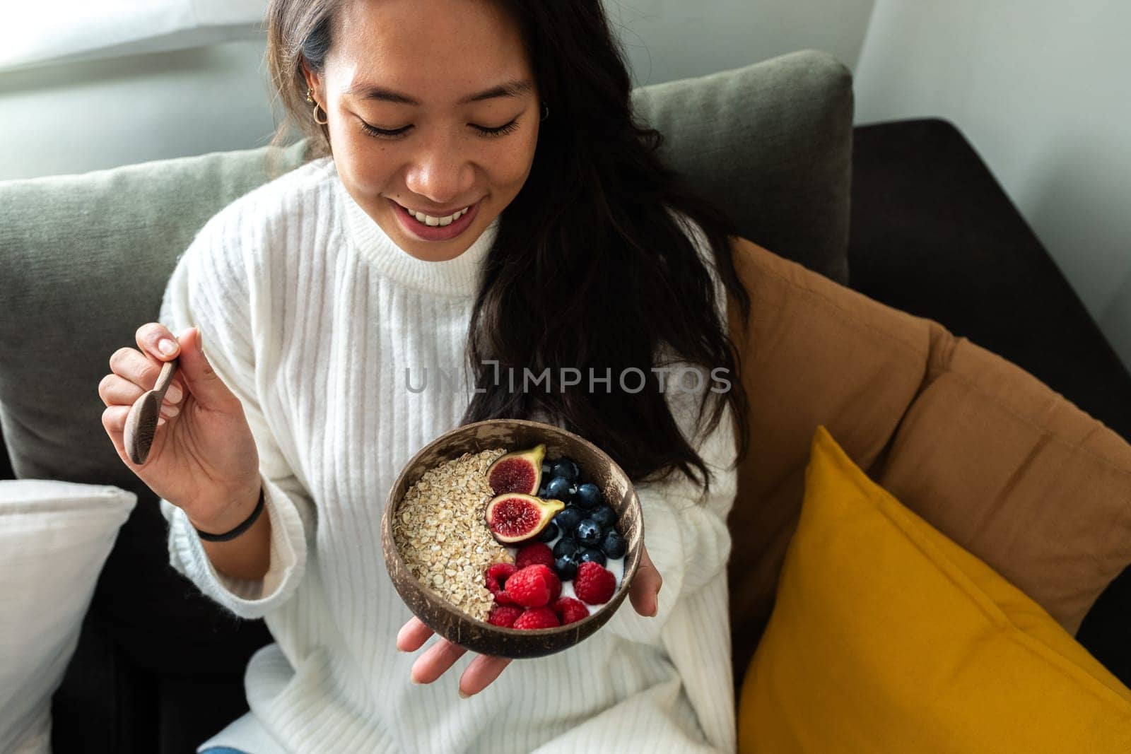 Happy young Asian woman smiling eating bowl of healthy breakfast oats with fruit sitting on the sofa by Hoverstock