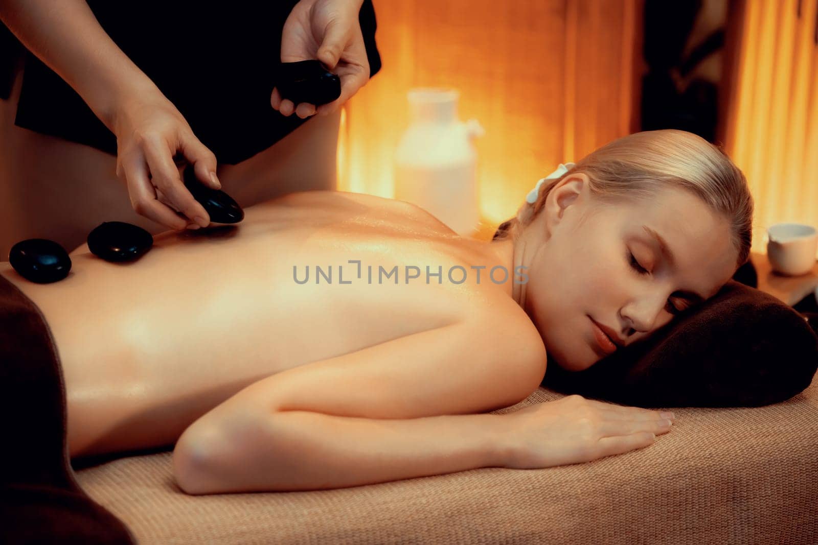 Hot stone massage at spa salon in luxury resort with warm candle light, blissful woman customer enjoying spa basalt stone massage glide over body with soothing warmth. Quiescent