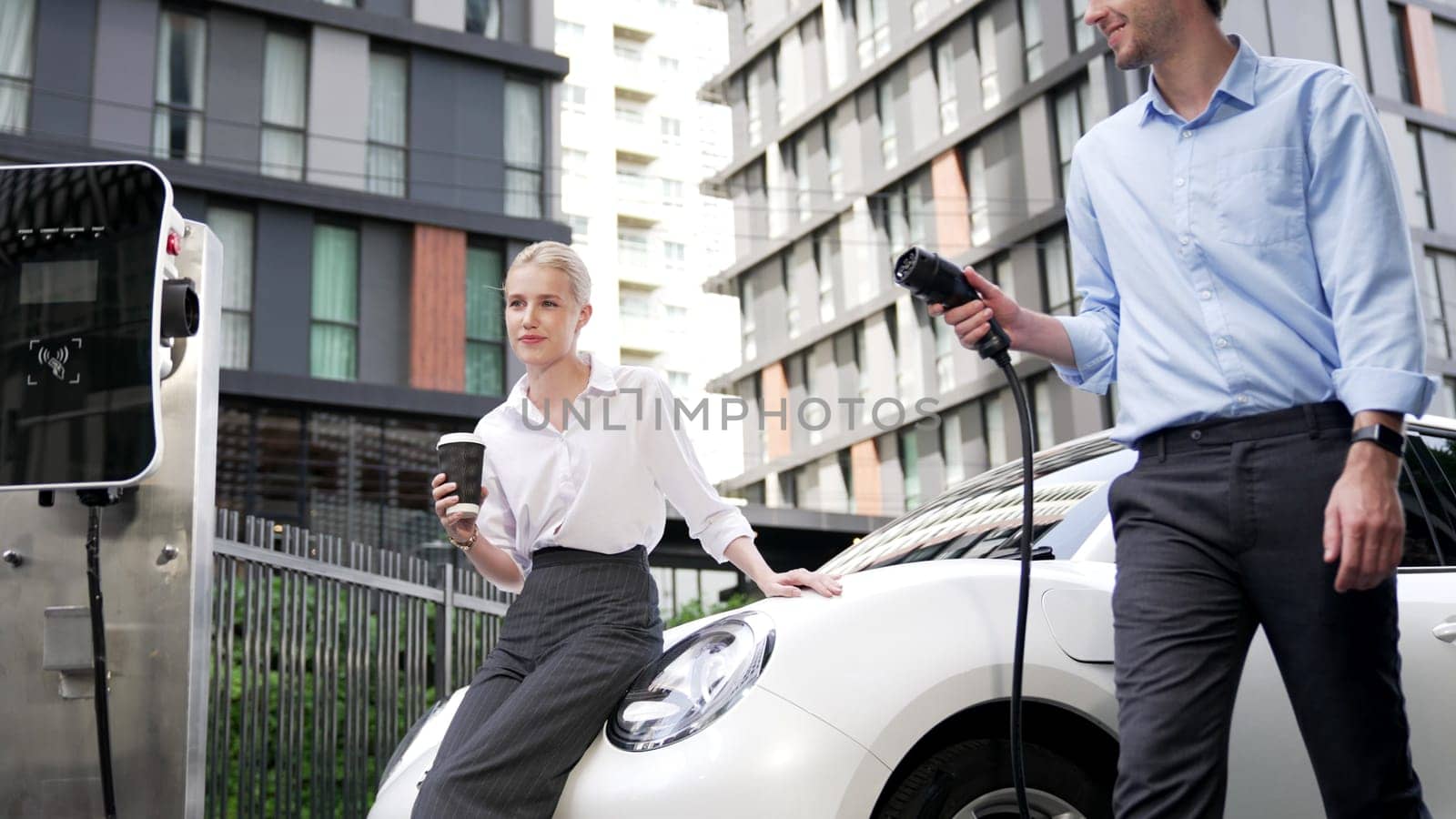 Businessman and businesswoman with progressive ambition unplug power cable from electric vehicle, standing on charging station with power cable plug and renewable energy-powered electric vehicle.