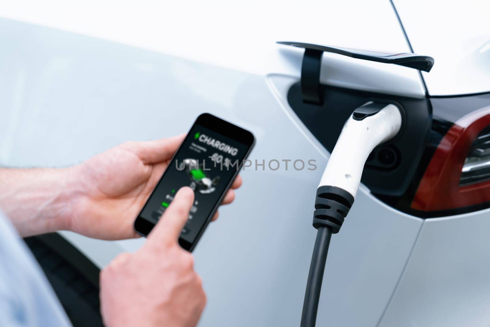 Modern eco-friendly man recharging electric vehicle from EV charging station, using Innovative EV technology utilization for tracking energy usage to optimize battery charging on smartphone.Synchronos