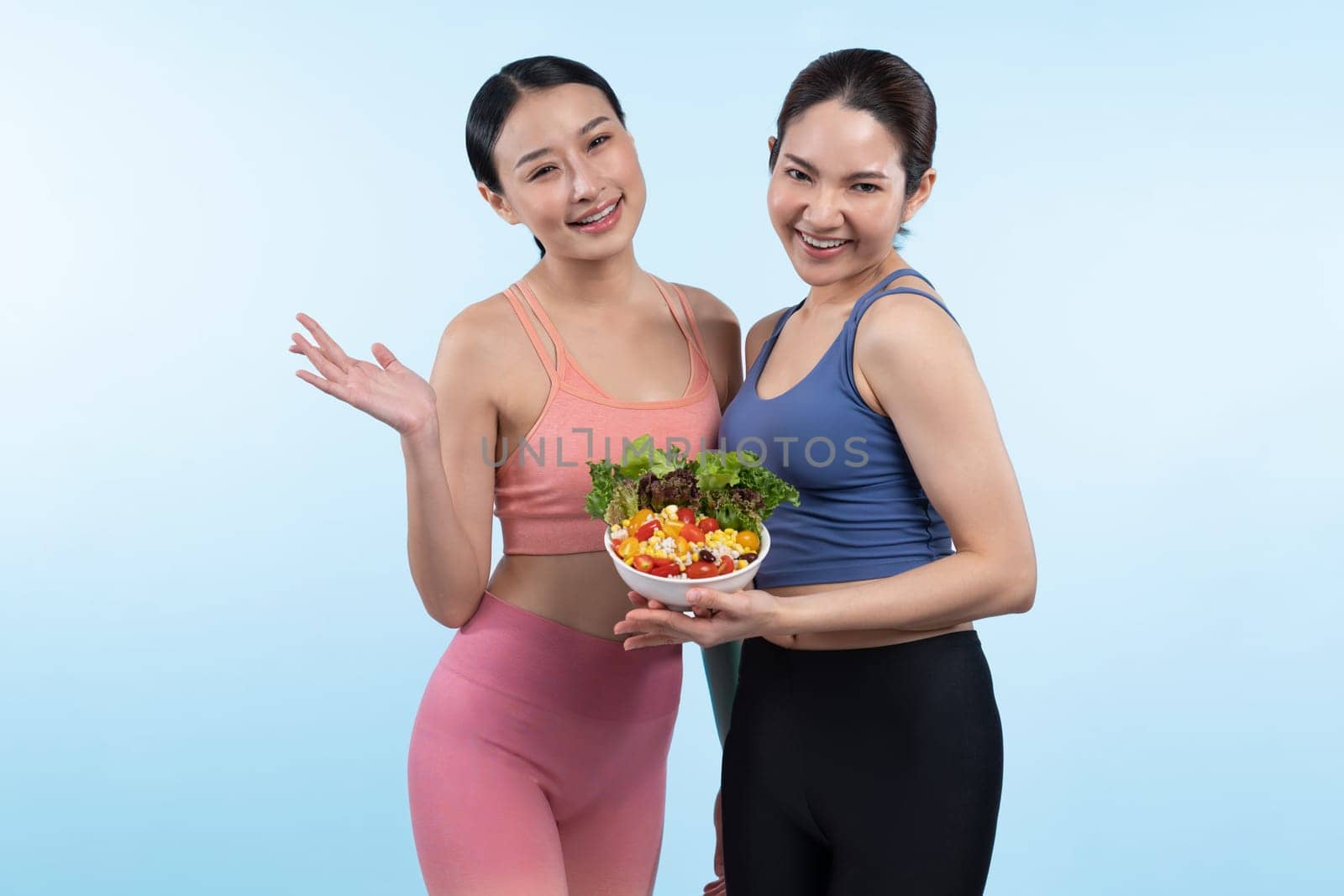 Two young sporty Asian women in sportswear holding salad bowl fill with fruit and vegetable. Natural youthful and fit body lifestyle people with balance nutrition on isolated background. Vigorous