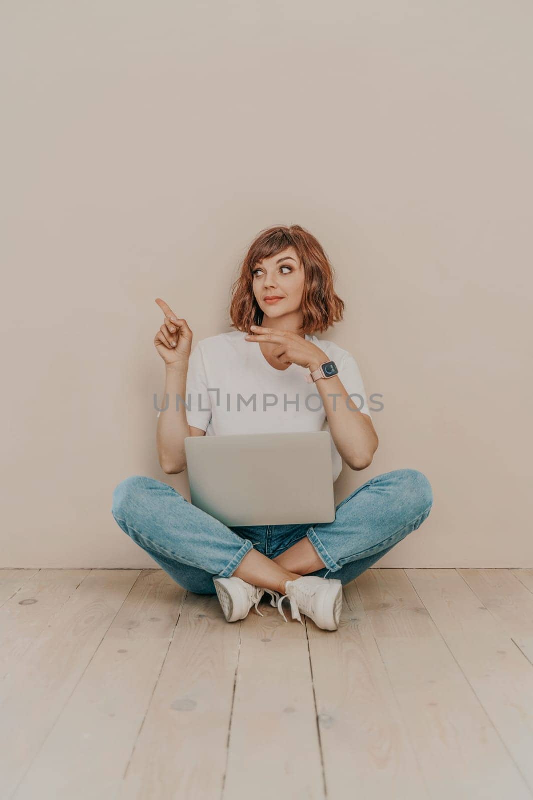 A brunette sits on the floor with a laptop on a beige wall background. She is wearing a white T-shirt, jeans and white sneakers. by Matiunina