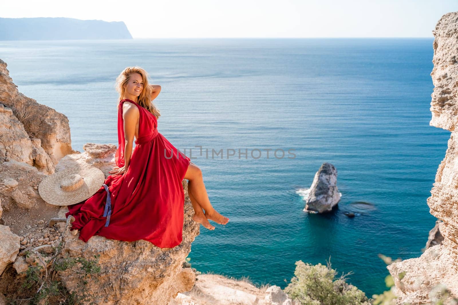 A girl with flowing hair in a long red dress sits on a rock above the sea. The stone can be seen in the sea. by Matiunina