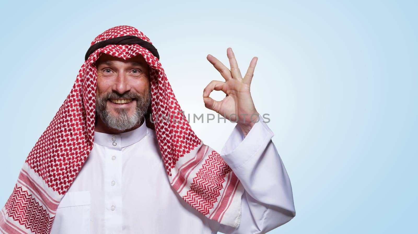 Cheerful and confident Arab man smiles warmly and shows OK sign against light blue background, leaving space for text. Approval, and contentment, making versatile choice for various messaging needs. High quality photo