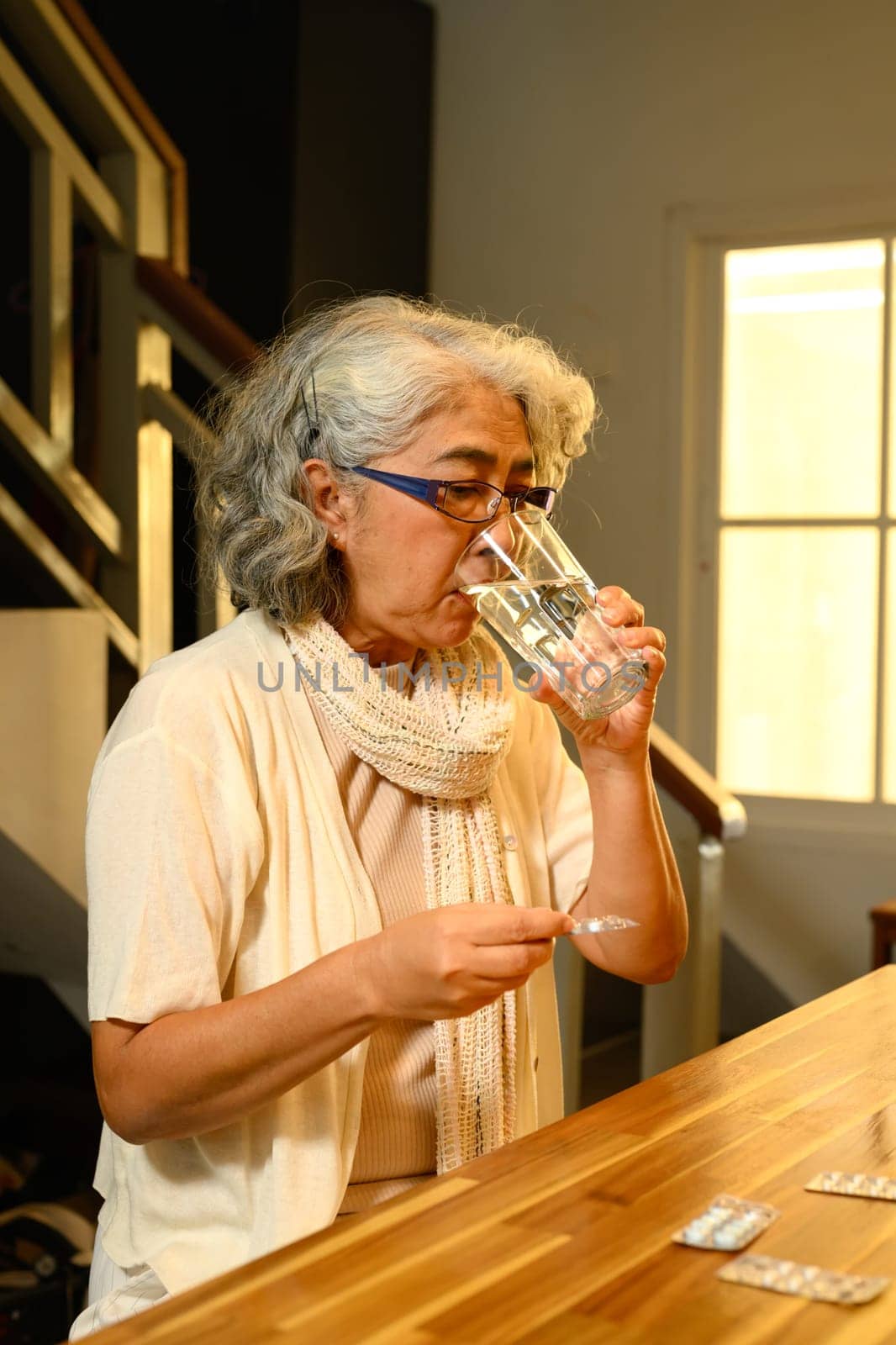 Mature woman taking her medication with a glass of water at kitchen table. Health care concept.