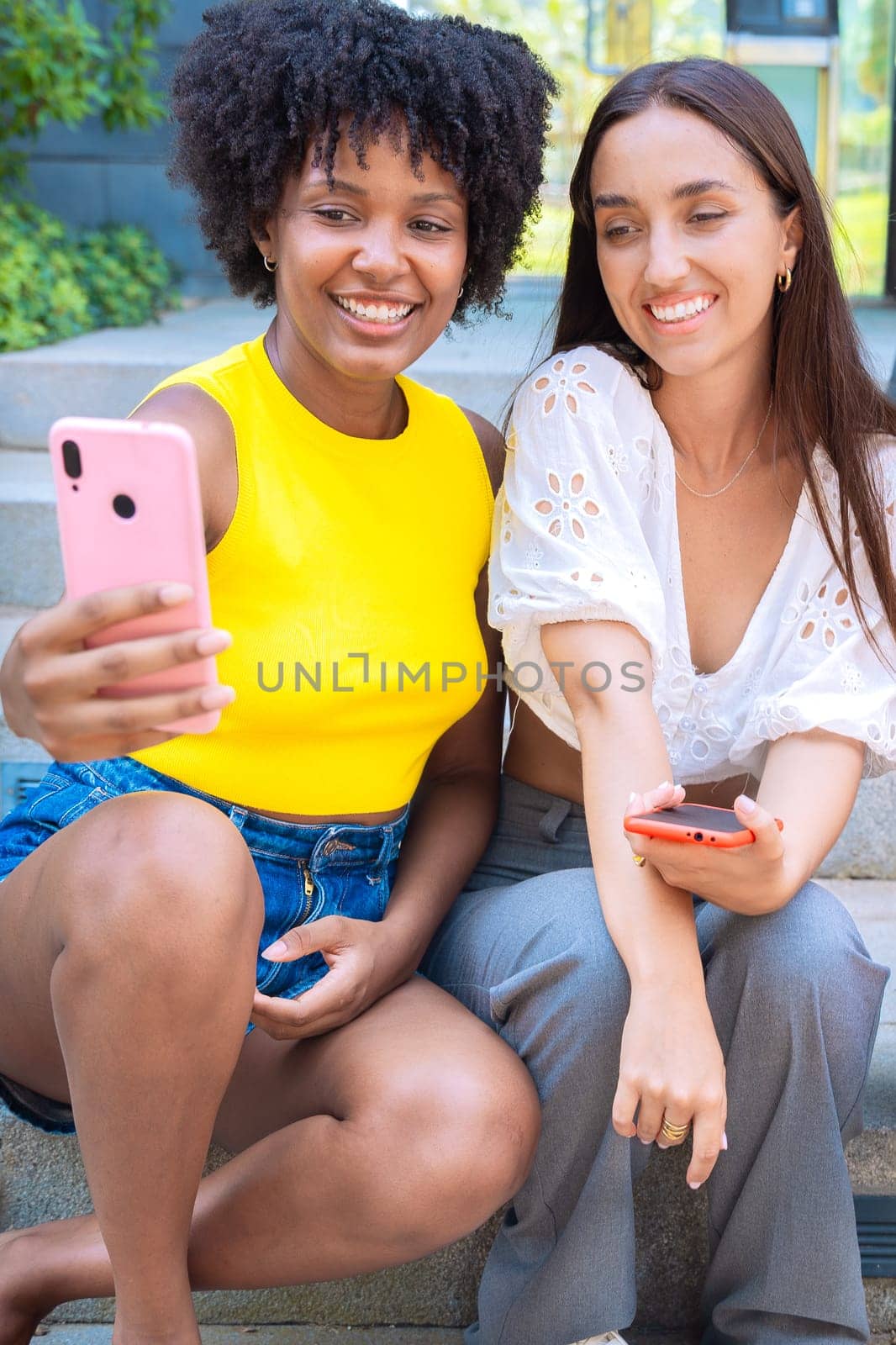 Two young women sitting outdoors, smiling, and taking a playful selfie together. Perfect for concepts like friendship, happiness, social media, and outdoor lifestyle.