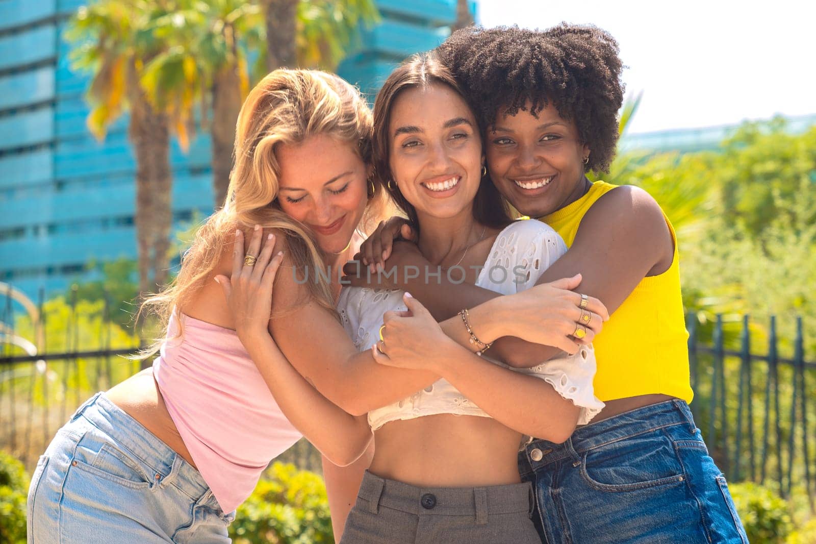 Three happy young women share a warm embrace while smiling outdoors. Their expressions reflect joy, friendship, and affection in a relaxed urban setting with palm trees and modern buildings.