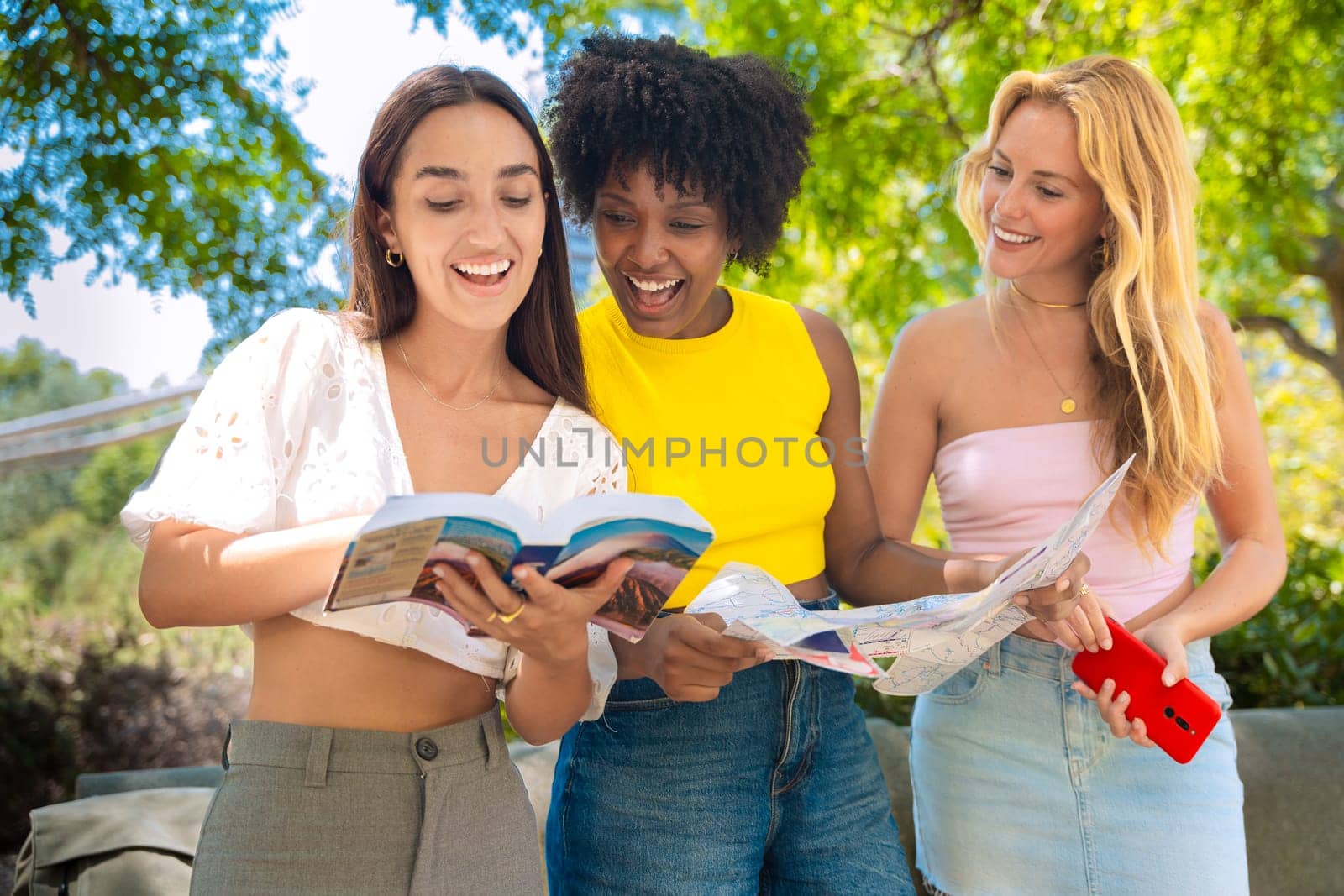 Three happy young women stand outdoors, looking at maps and a travel guide. They smile while planning their trip, enjoying a fun adventure in a sunny urban setting with trees and buildings.