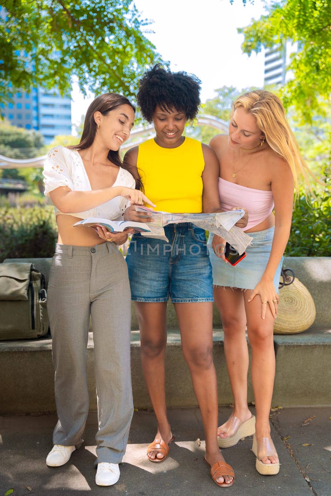 Three happy young women stand outdoors, looking at maps and a travel guide. They smile while planning their trip, enjoying a fun adventure in a sunny urban setting with trees and buildings.