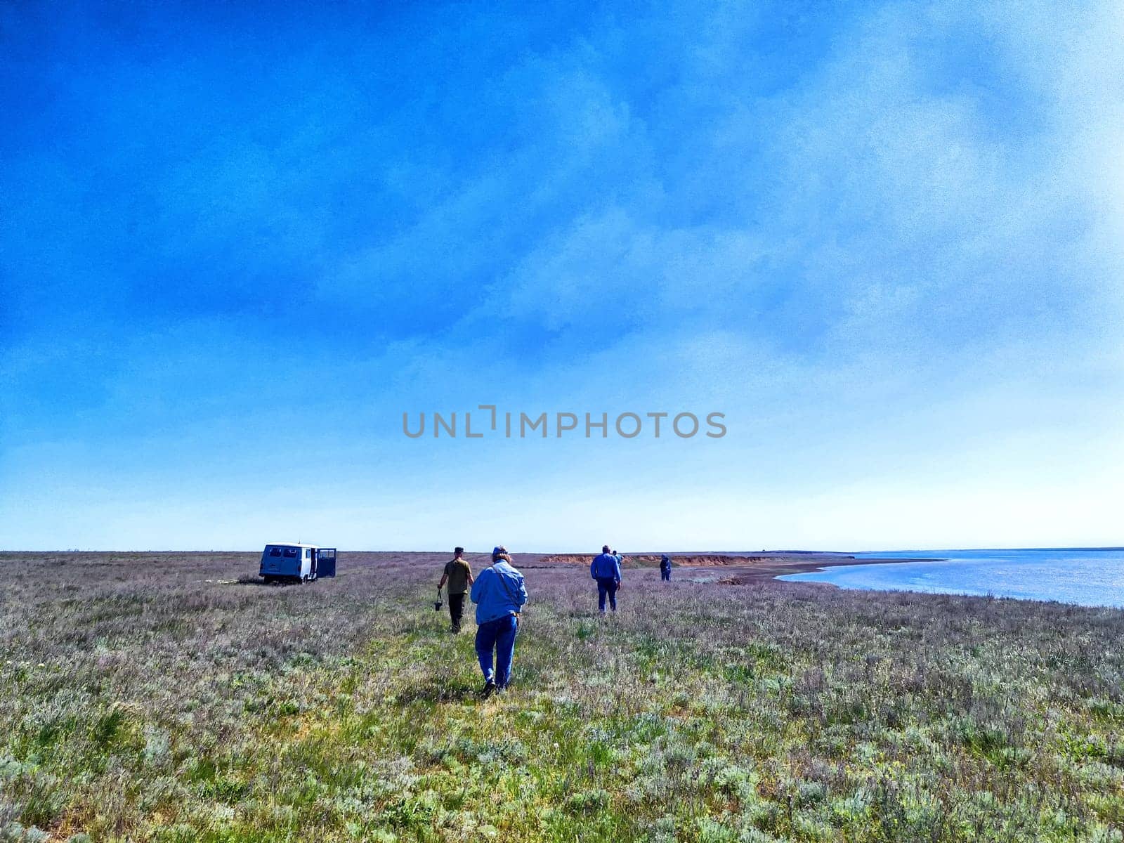 A group of individuals strolls across a sprawling grassland adjacent to a calm shoreline under a bright blue sky.