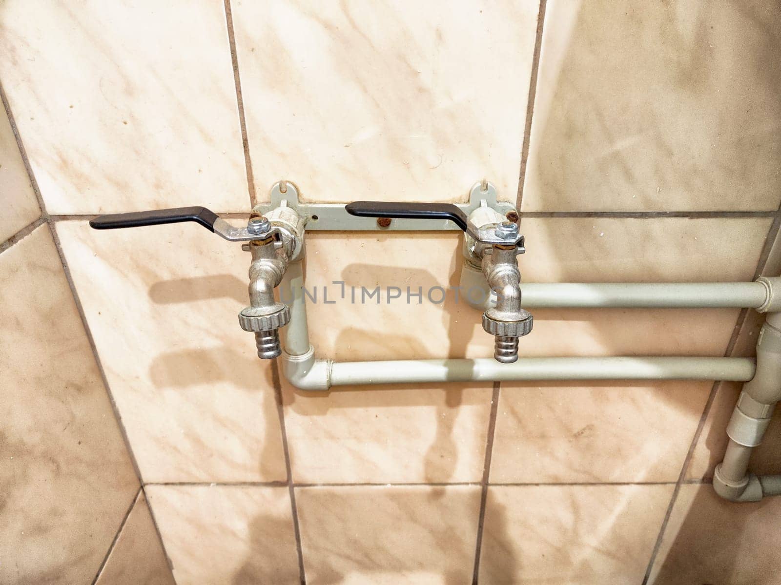 Two metallic plumbing taps are mounted on a tiled wall, connected by pipes, and ready for use in a utility space.