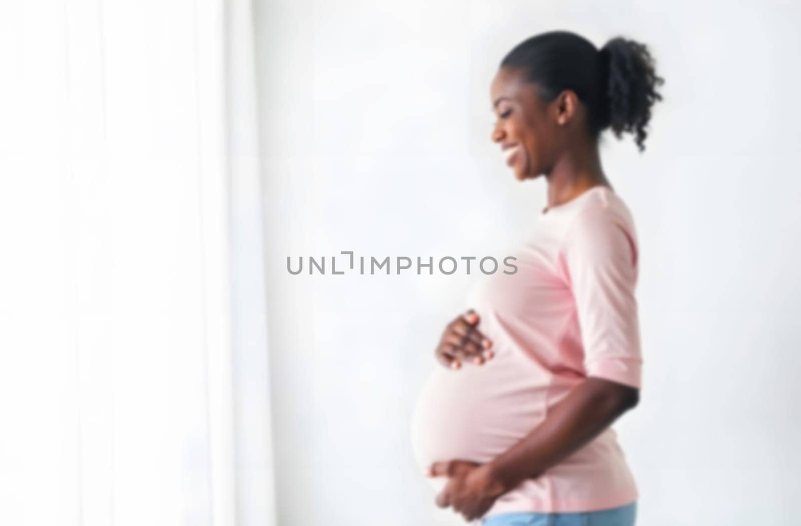 Happy expectant mother stands by a window, cradling her baby bump with a joyful expression in a softly lit room.. Blurred picture