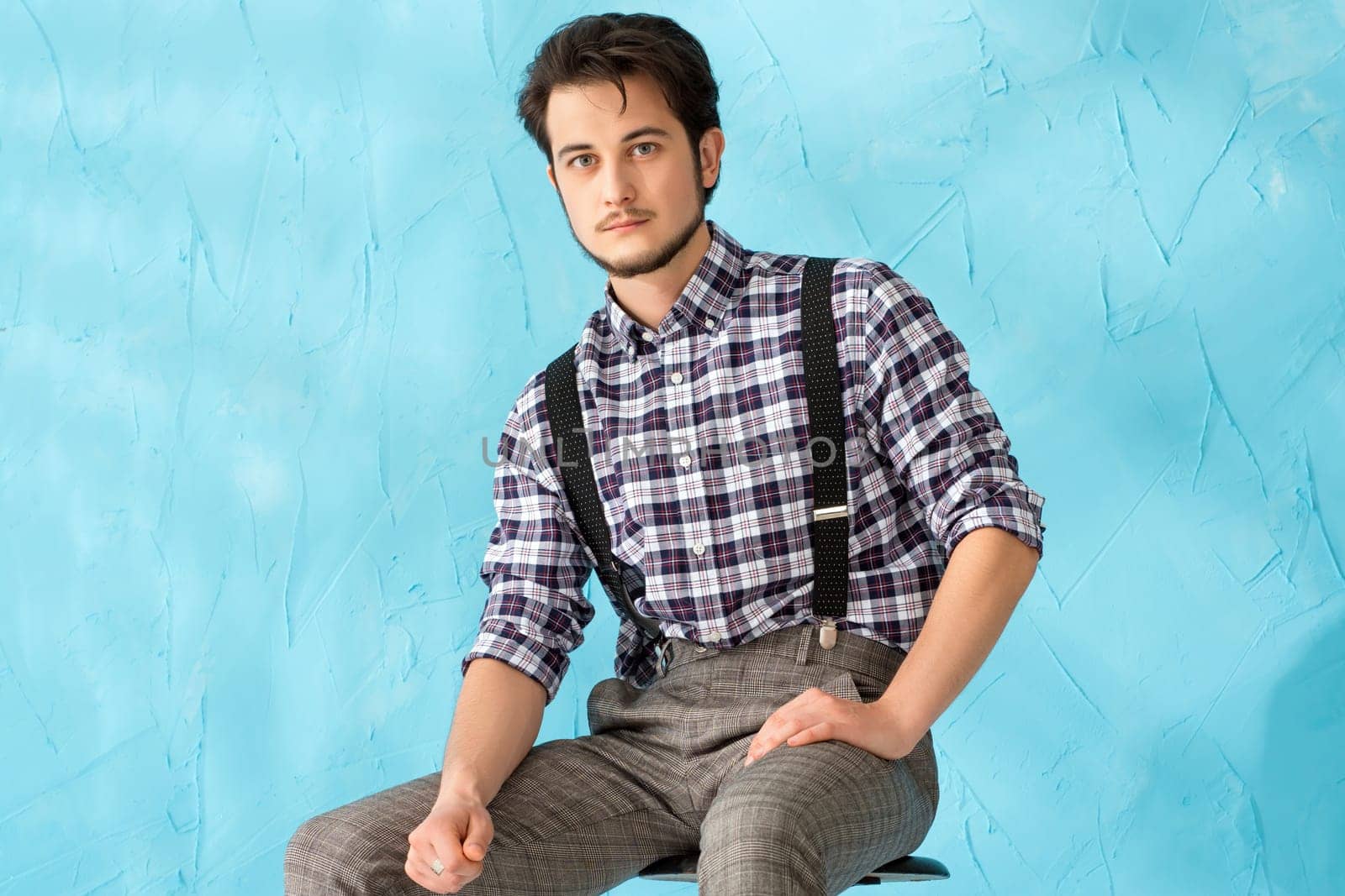 A young man in a plaid shirt and suspenders sits confidently on a chair, showcasing a relaxed fashion style against a blue backdrop.