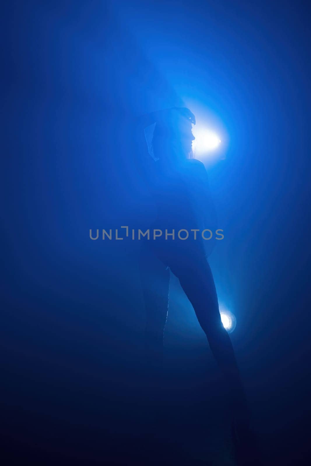 A figure moves through dense blue fog, illuminated by bright lights, creating an ethereal atmosphere in an unseen location.