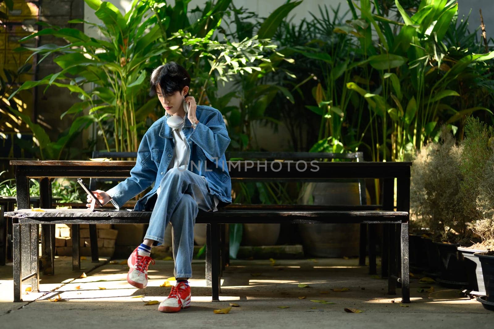 Young creative man wearing headphones sitting on a bench in a lush outdoor and using digital tablet.