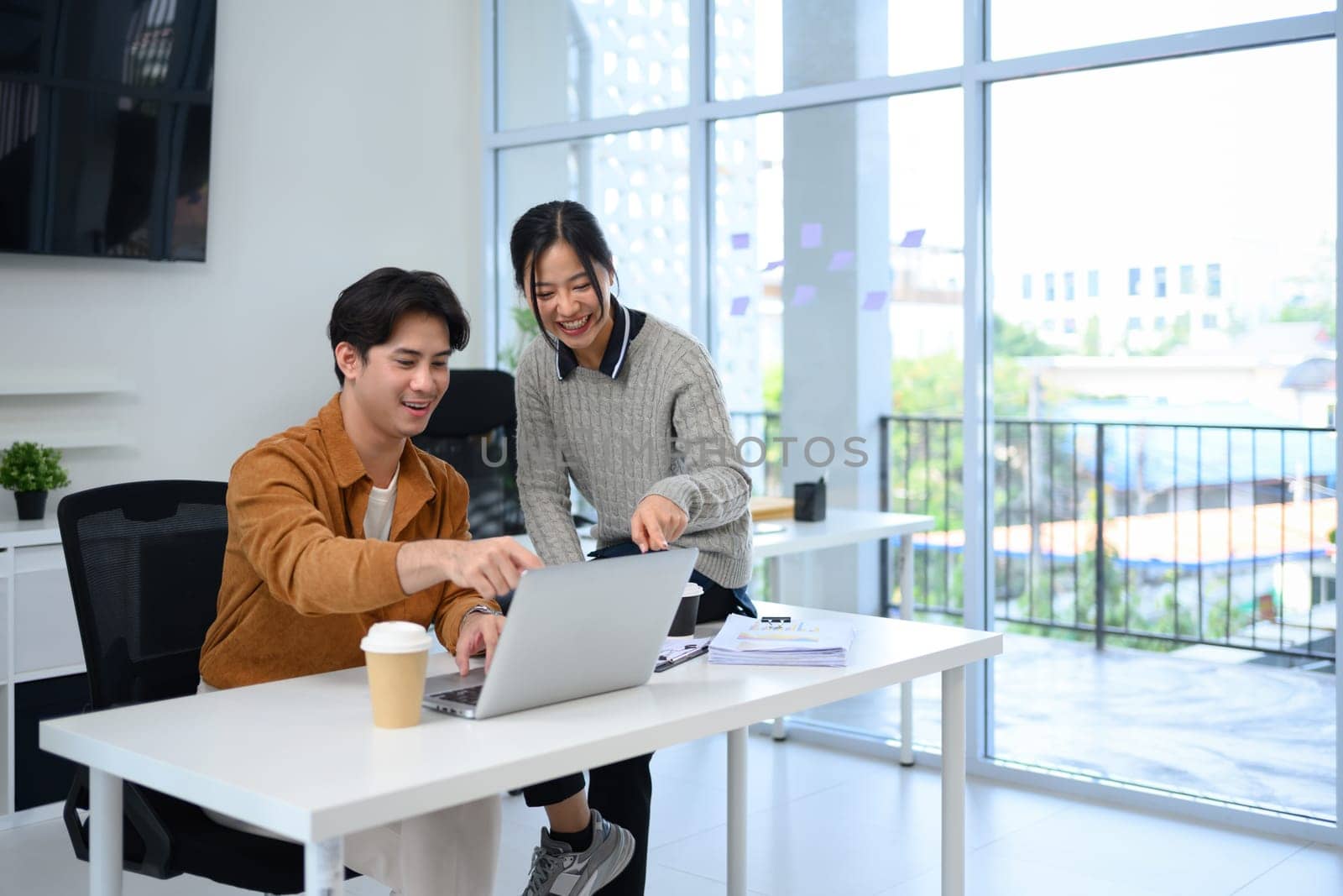 Two smiling coworkers reviewing project details on laptop at modern workplace.