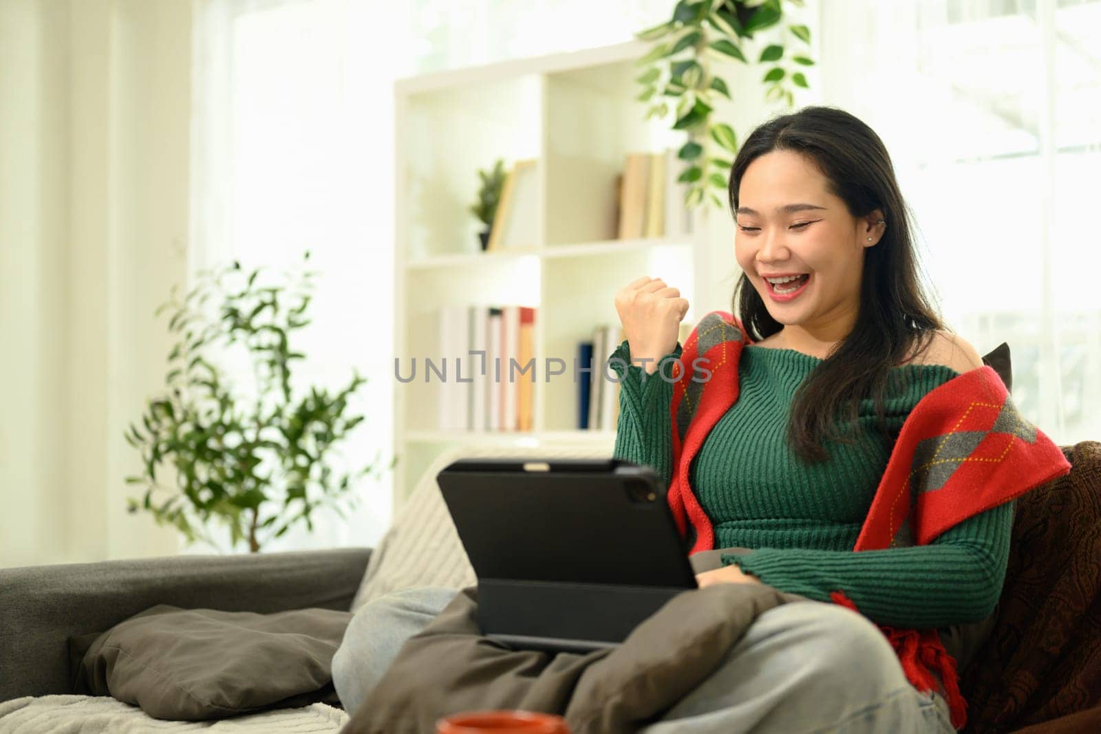 Happy woman in a green sweater celebrating while using digital tablet on her lap.