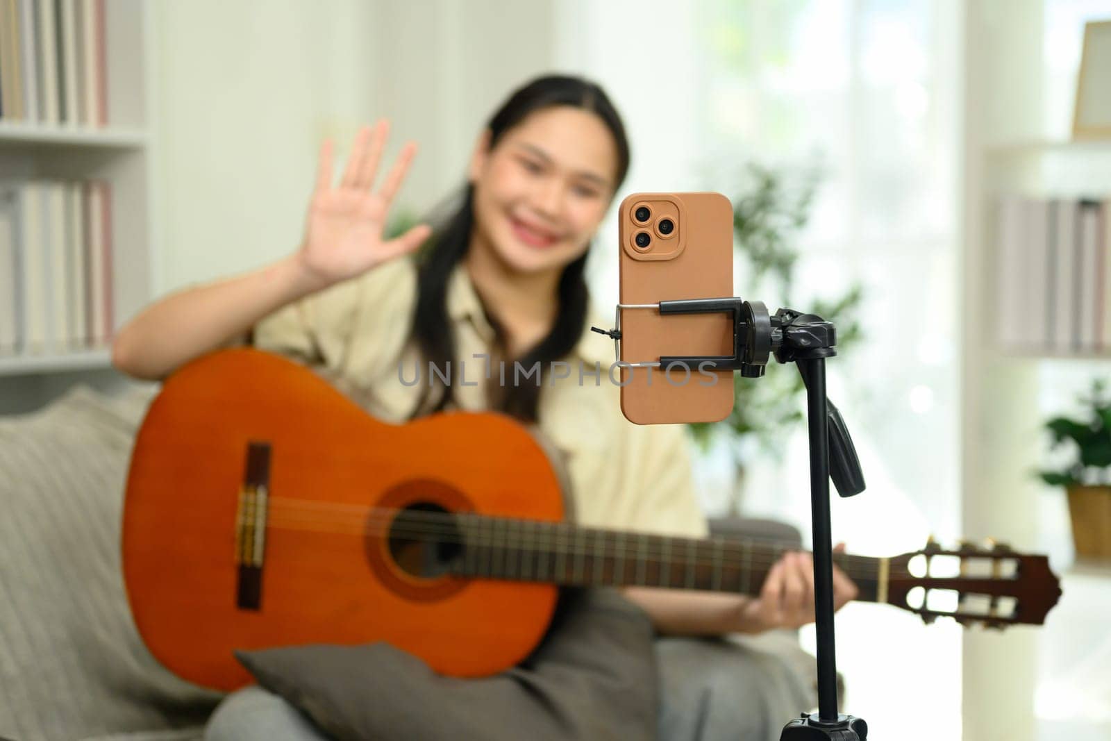 Cheerful woman playing guitar and waving at the camera during a live stream or video recording session.