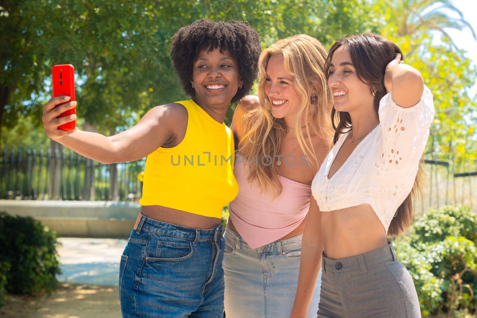 Three young women sitting outdoors, smiling, and taking a playful selfie together. Perfect for concepts like friendship, happiness, social media, and outdoor lifestyle.