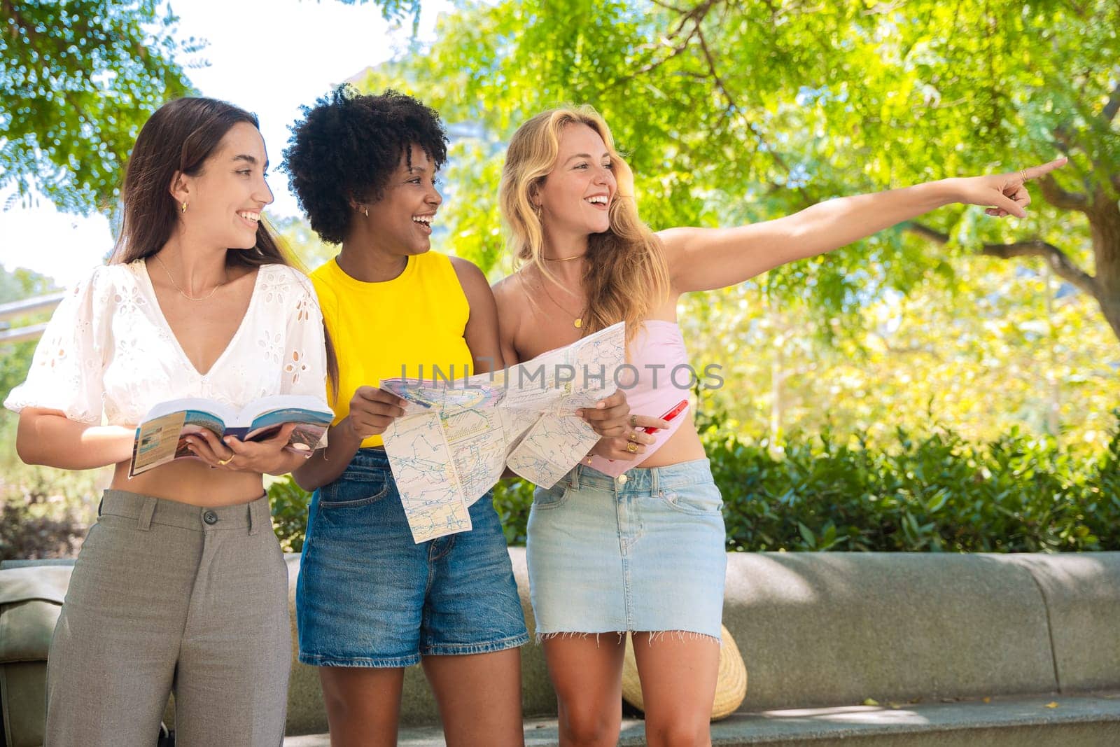 Three cheerful young women stand outdoors, holding maps and a travel guide while pointing at something. They smile excitedly, enjoying their adventure in a sunny urban setting with trees and buildings