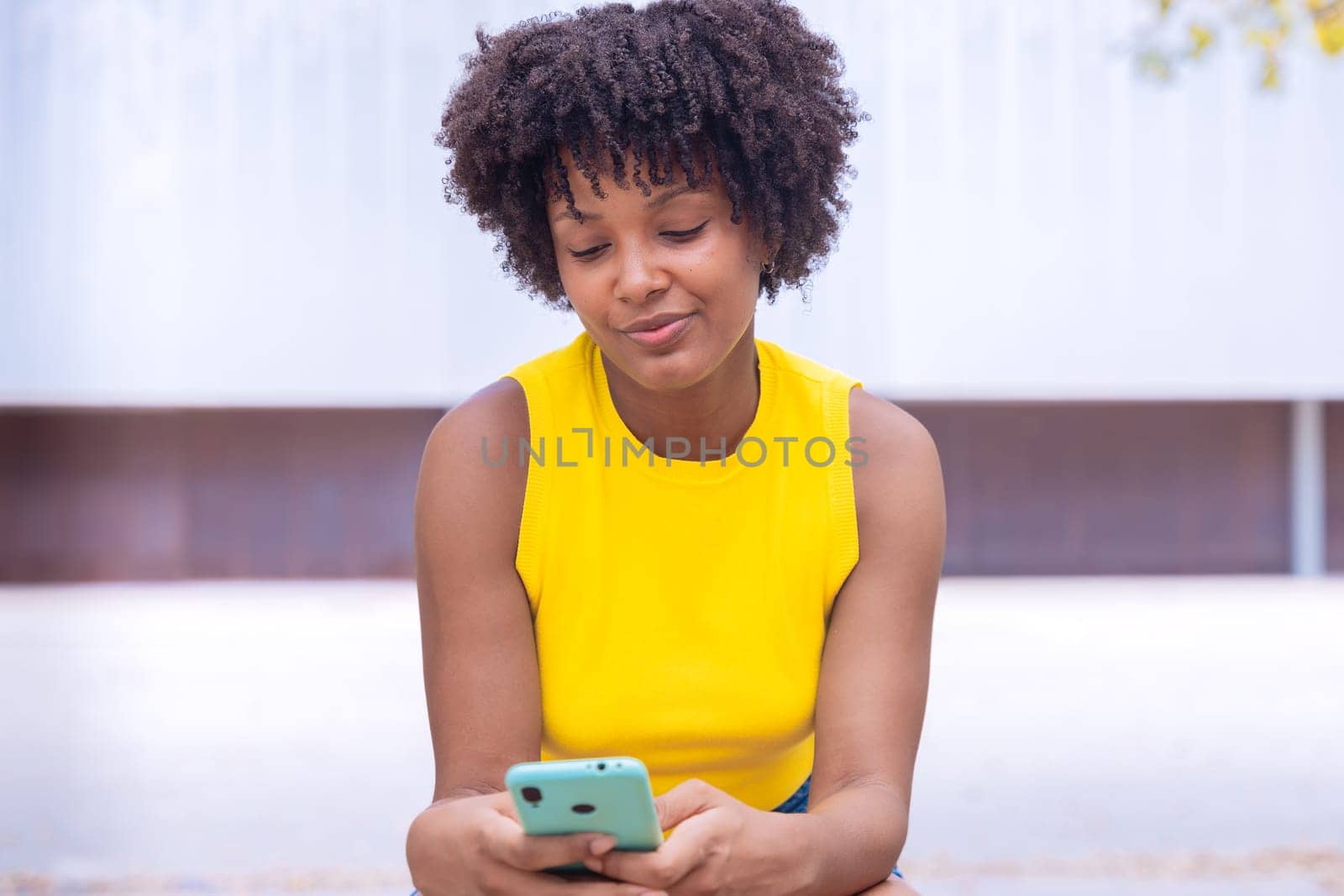 A young woman with natural curly hair wearing a yellow top rests her face on her hand while holding a smartphone. Calm and happy moment captured in a bright outdoor setting.