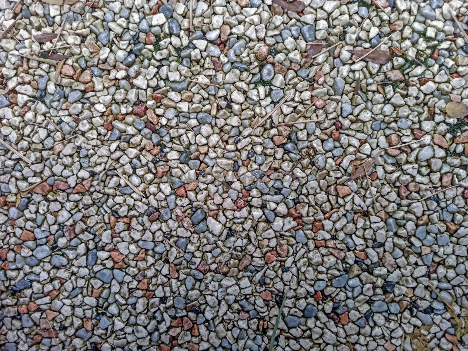 A variety of small pebbles in different colors covers the ground in a garden. These stones create an interesting and colorful texture under the warm sunlight. Natural elements surround the pebbles.