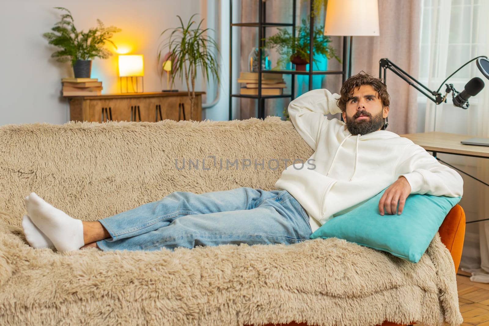 Tired Indian handsome young man enjoy relaxing on home sofa in living room. Happy guy lying on couch resting napping after hard working day, closed his eyes, put his hand behind his head, stress free.
