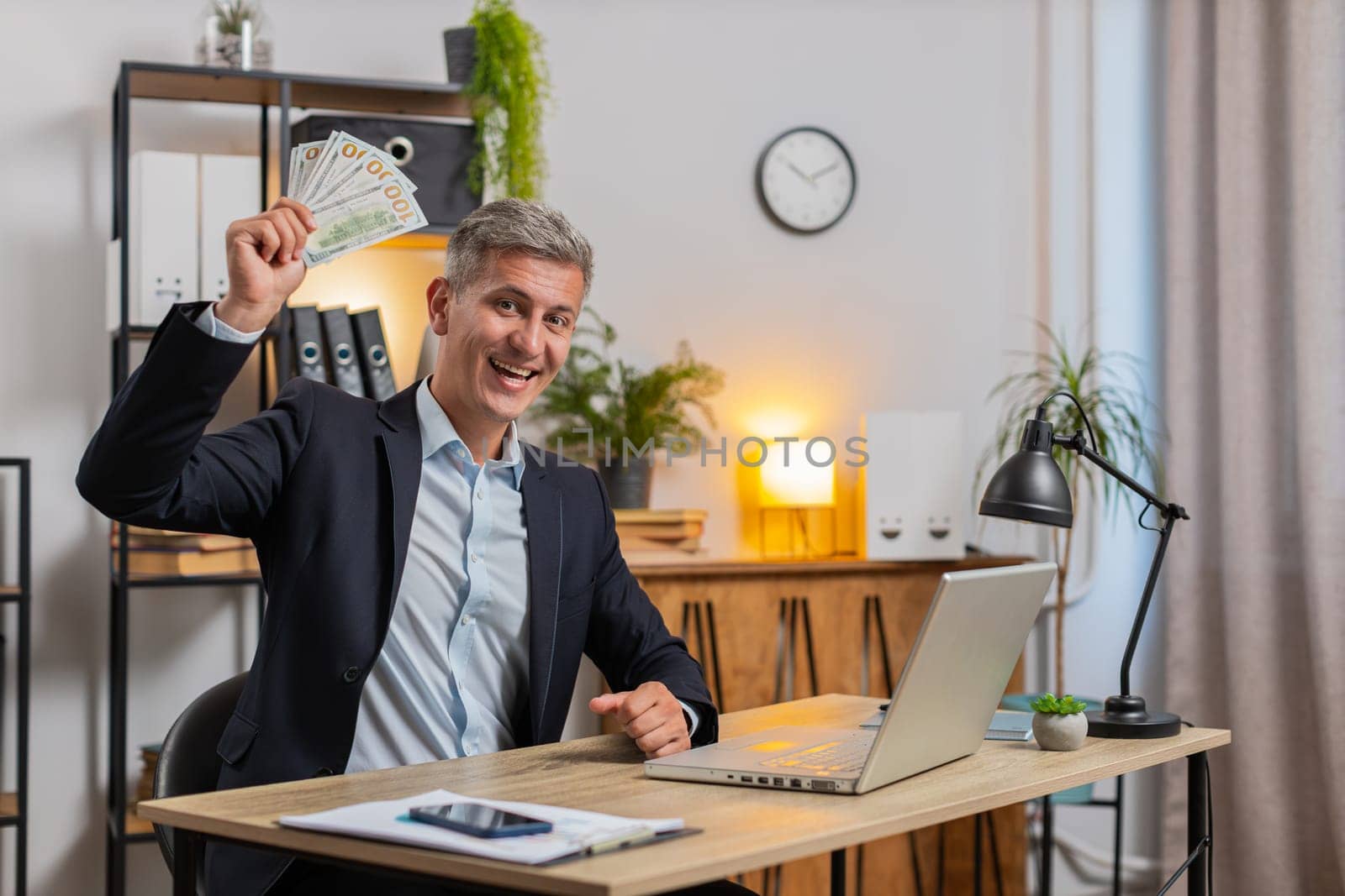 Caucasian young businessman holding fan of cash money dollar banknotes celebrate, success career, lottery jackpot game winner, big income, wealth at home office workplace desk. Remote distant working.