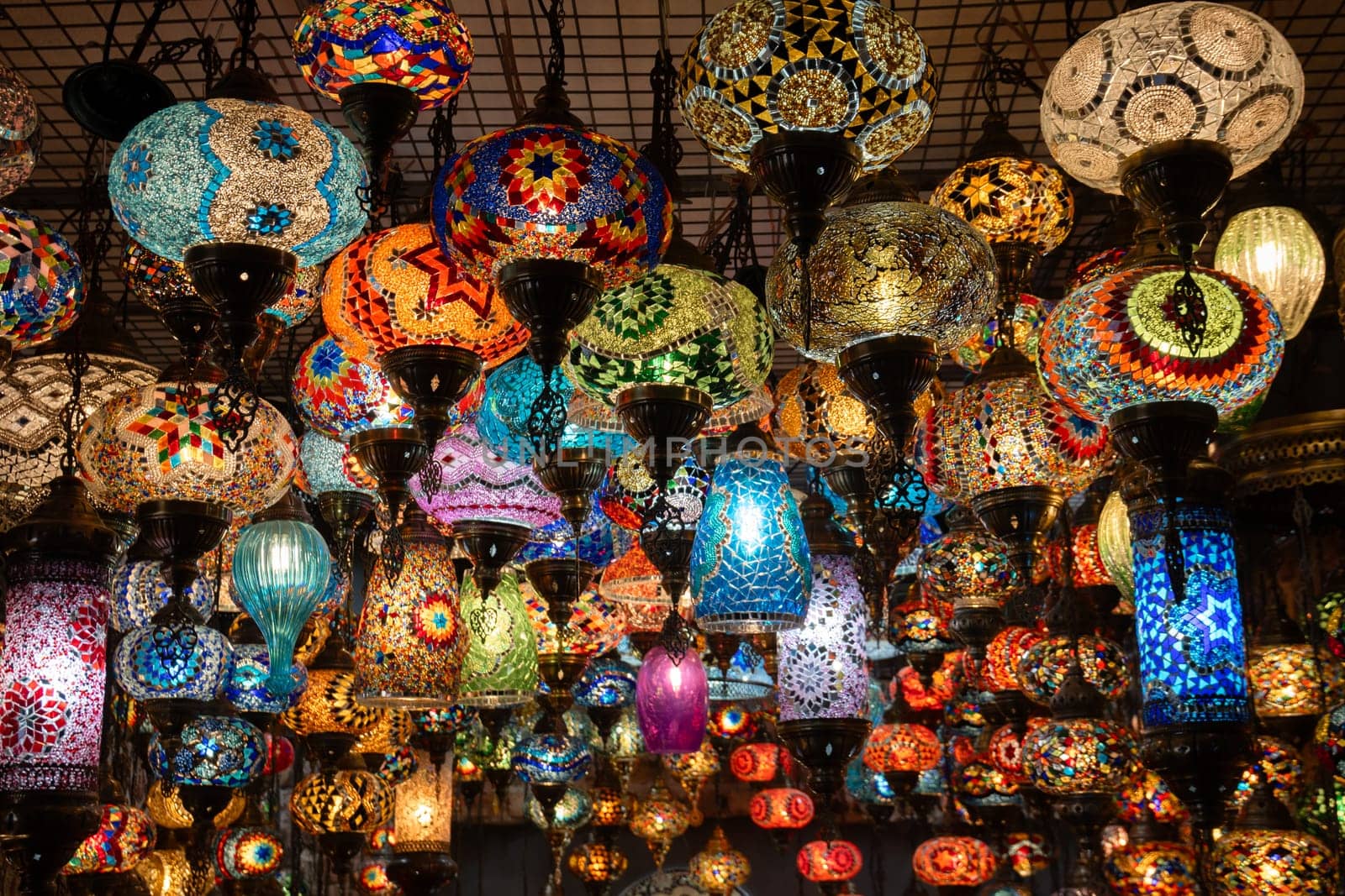 A dense array of colorful, hand-painted glass lamps hangs from a ceiling, illuminated, showcasing intricate designs and varied shapes in a vibrant display.
